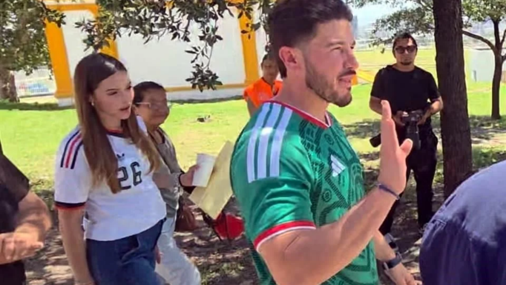 Costo-del-viaje-de-samuel - Tu Colonia Regia Man in a Green Sports Jersey Raises His Hand While Walking in a Sunny Park, with a Woman in a White Jersey (number 26) and Others Nearby in the Background. - Tu Colonia Regia