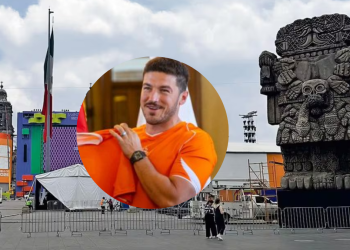 Outdoor Festival Scene with a Tall Flag, Colorful Stage Structures, and a Large Stone Aztec Statue in the Background; a Man in an Orange Shirt Appears in a Circular Overlay at the Center. - Tu Colonia Regia