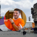 Fifa-fanfest - Tu Colonia Regia Outdoor Festival Scene with a Tall Flag, Colorful Stage Structures, and a Large Stone Aztec Statue in the Background; a Man in an Orange Shirt Appears in a Circular Overlay at the Center. - Tu Colonia Regia
