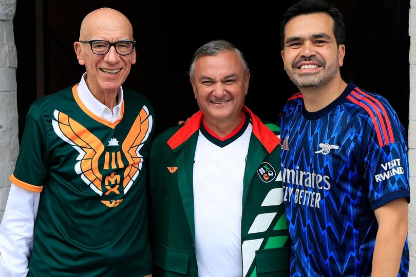 Image - Tu Colonia Regia Three Men Stand Shoulder to Shoulder and Smile, Each Wearing a Different Soccer Jersey (green with Eagle Emblem, Green Jacket, Blue Away Kit). - Tu Colonia Regia