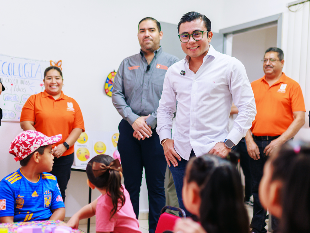 Centros_comunitarios_nl - Tu Colonia Regia Man in a White Shirt and Glasses Smiles at Children Seated Around a Classroom Table, with Volunteers Standing in the Background. - Tu Colonia Regia