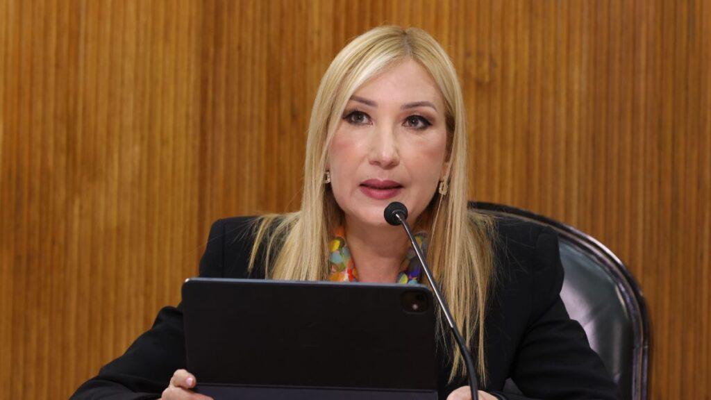 Secretaria-de-salud-pide-donaciones - Tu Colonia Regia Blonde Woman in a Black Blazer Speaks into a Microphone at a Formal Meeting, Seated Behind a Tablet in Front of a Wooden Panel Backdrop. - Tu Colonia Regia