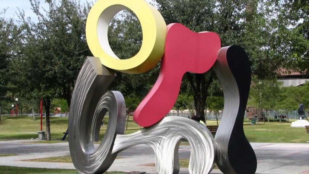 Brad-howe-escultura-cineteca - Tu Colonia Regia Outdoor Abstract Metal Sculpture with Looping Shapes in Yellow, Pink, Black and Silver on a Park Lawn - Tu Colonia Regia