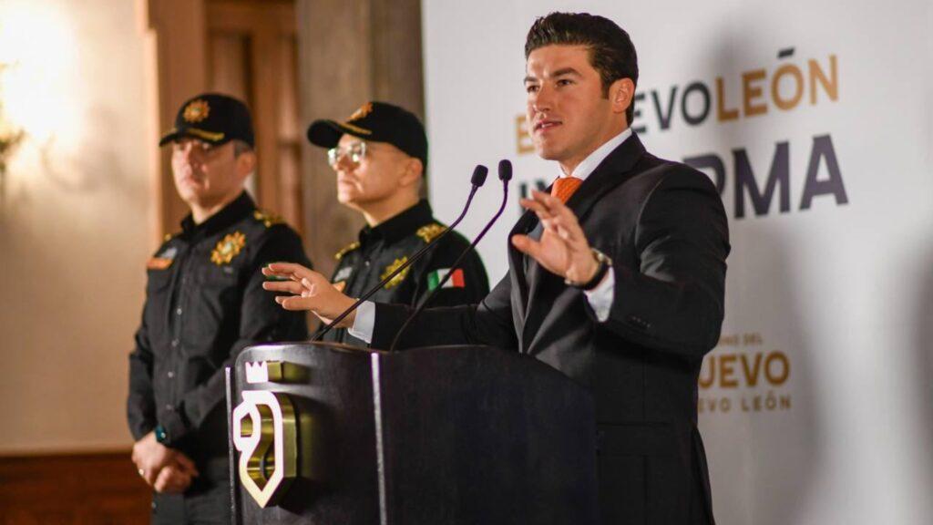 a Man in a Dark Suit Speaks at a Podium with Two Uniformed Officers Standing Behind Him, Near a Banner. - Tu Colonia Regia